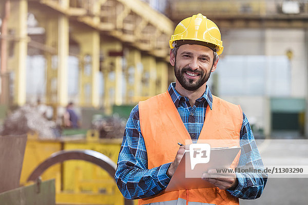 Portrait lächelnder Stahlarbeiter mit Klemmbrett in der Fabrik