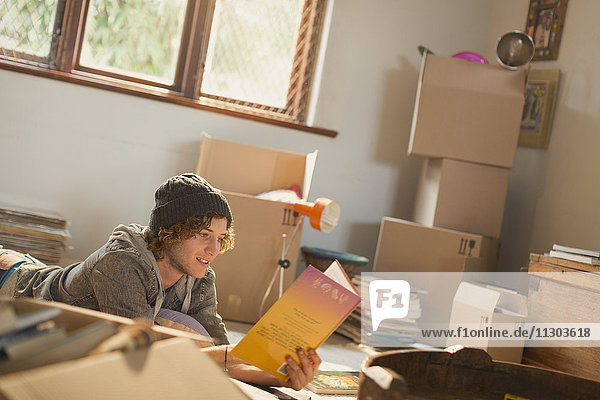 Young man reading book surrounded by moving boxes in apartment