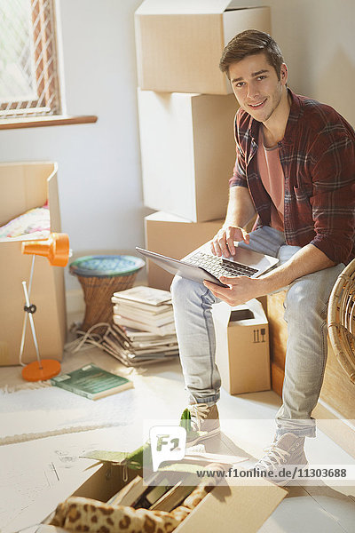 Porträt eines lächelnden jungen Mannes  der einen Laptop benutzt  umgeben von Umzugskartons in einer neuen Wohnung