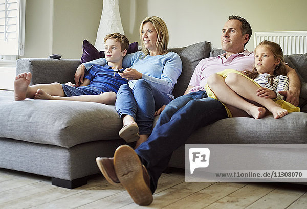 A family of four people  parents and a girl and boy  seated on the sofa together  watching television.