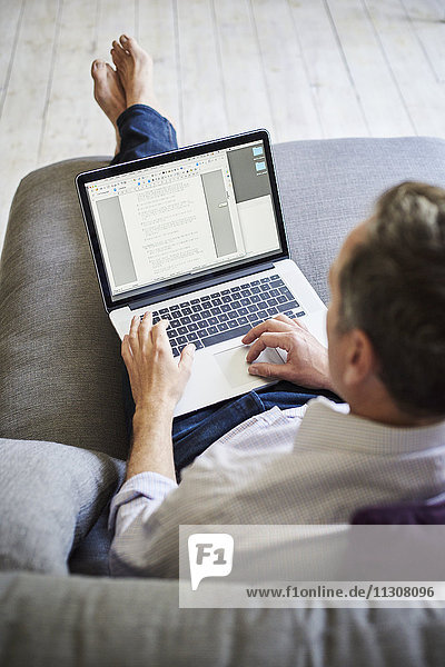 A man seated on a sofa  using a laptop computer  over the shoulder view.