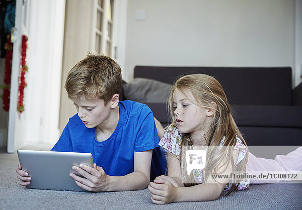 Two children lying on their stomachs sharing a digital tablet watching the screen.