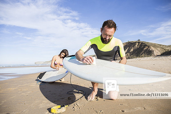 Couple putting paraffin on their surfboards on the beach