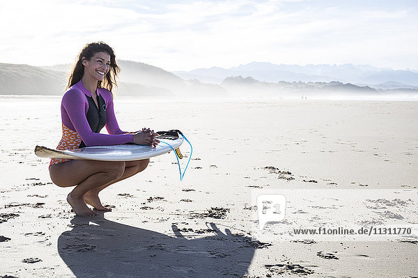 Smiling woman with surfboard crouching on the beach