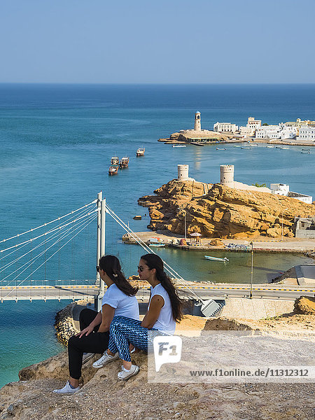 Oman  Ash Sharqiyah  Ad Daffah  Rückansicht von zwei Frauen auf einem Felsen vor der Hafenstadt Sur