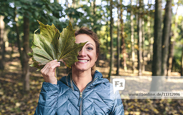 Grinsende Frau bedeckt das Auge mit Herbstblatt im Wald
