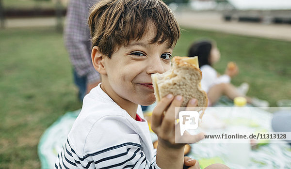 Porträt eines lächelnden Jungen  der ein Sandwich mit seiner Familie im Hintergrund hält.