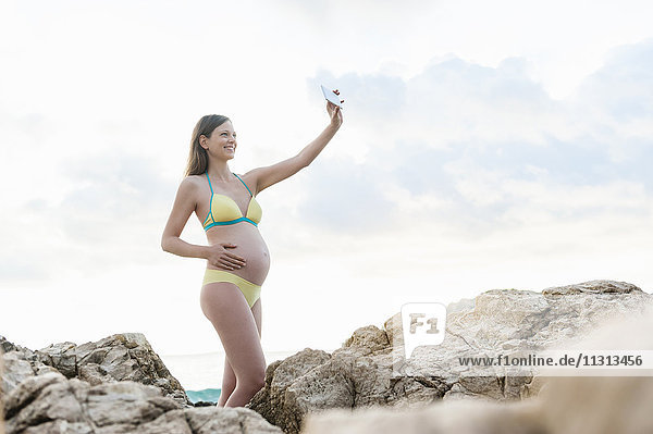 Schwangere Frau zwischen Felsen am Meer mit einem Selfie