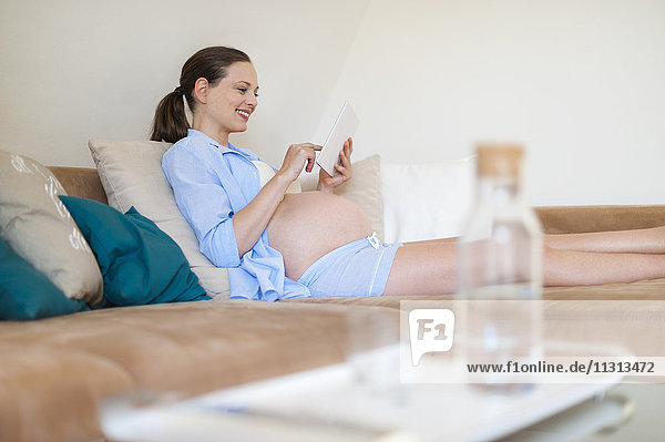 Lächelnde schwangere Frau auf der Couch liegend mit Tablette