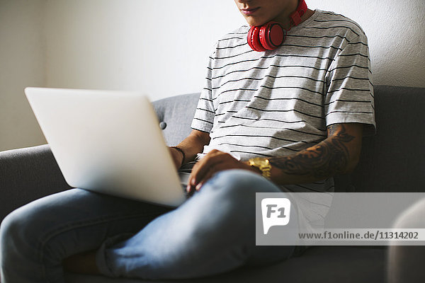Teenage boy with headphones sitting on armchair at home using laptop