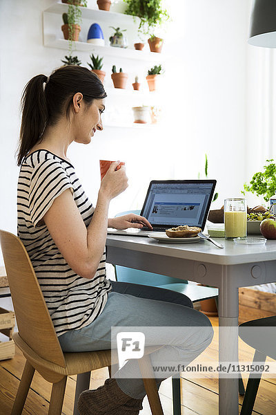 Lächelnde Frau zu Hause am Tisch sitzend mit Laptop