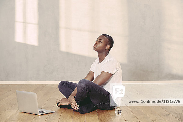 Tagträumender junger Mann auf dem Boden sitzend mit Laptop und Wasserglas
