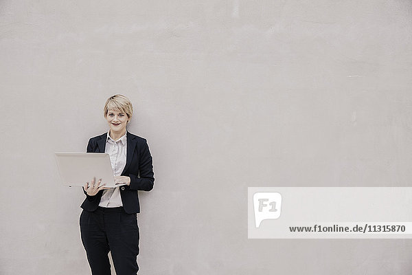 Lächelnde Geschäftsfrau mit Laptop vor der Betonwand stehend