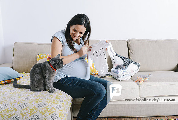 Schwangere Frau mit Katze  die auf der Couch sitzt und Babyschläfer ansieht.