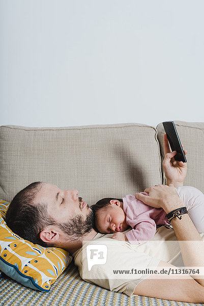 Vater auf der Couch liegend mit neugeborenem Mädchen über Smartphone