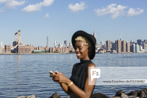 USA  New York City  Brooklyn  lächelnde junge Frau am East River mit Manhattan-Skyline im Hintergrund
