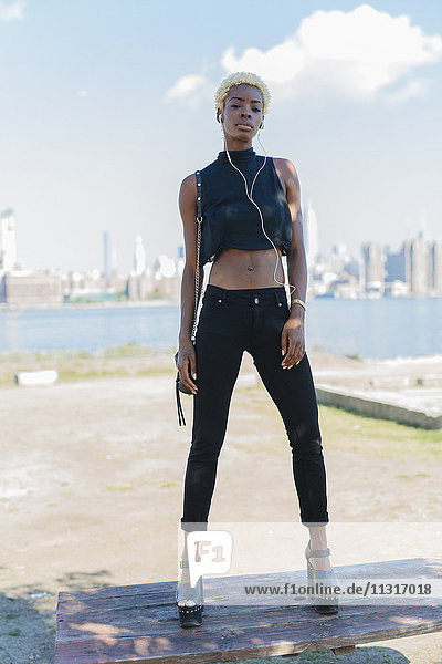 USA  New York City  Brooklyn  selbstbewusste junge Frau mit Ohrstöpseln auf Holztisch stehend