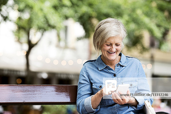 Lächelnde ältere Frau sitzt auf der Bank in der Stadt mit Smartphone