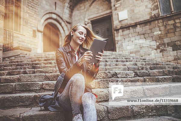Spain  Barcelona  smiling young woman sitting on stairs looking at tablet