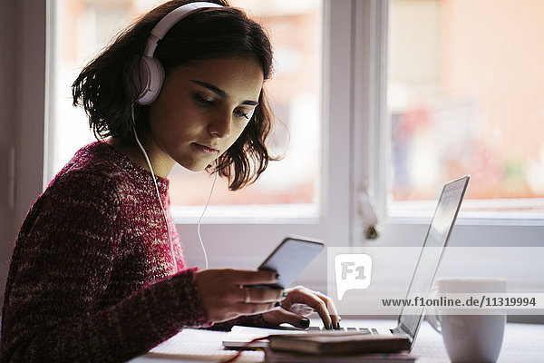 Junge Frau mit Laptop und Kopfhörer beim Blick aufs Handy