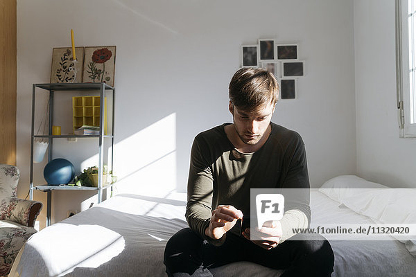 Young man sitting on bed looking at his smartphone