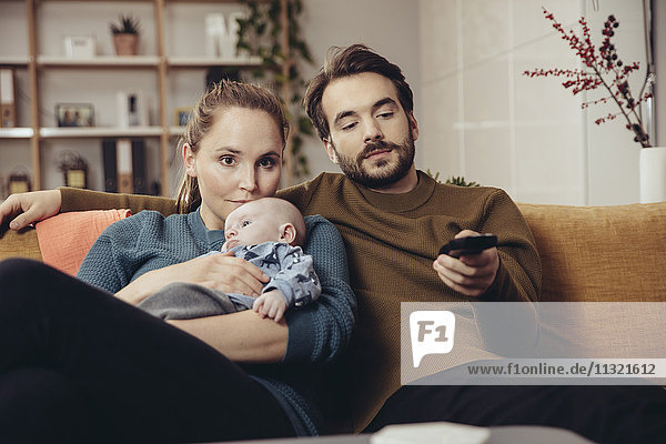 Father and mother sitting with baby on couch watching Tv