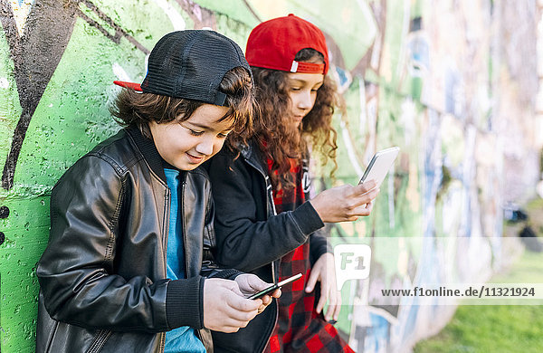 Two children leaning against graffiti wall using smartphones