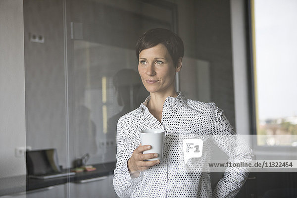 Lächelnde Frau mit einer Tasse Kaffee  die aus dem Fenster schaut.