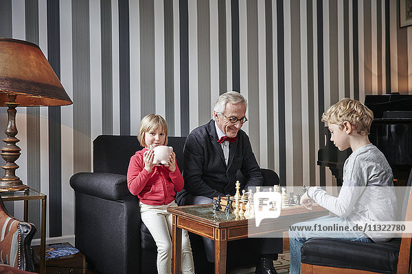 Grandfather and grandson playing chess in living room with girl sitting next to them