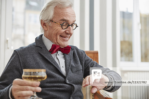 Lächelnder älterer Mann sitzt auf einem Stuhl und hält Champagnerglas und schaut auf smartwatch.
