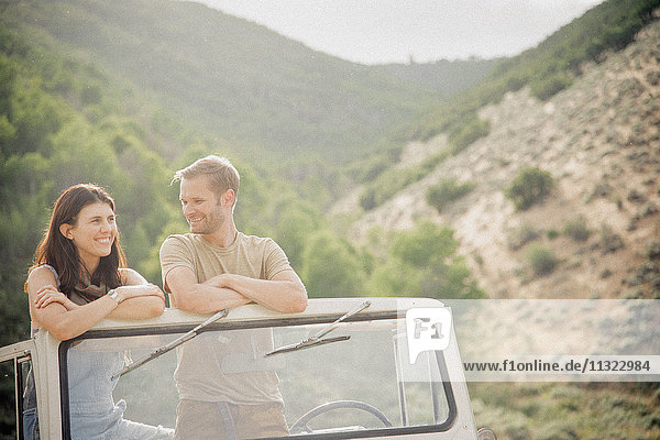 A couple on a road trip in the mountains standing side by side in a jeep looking around.