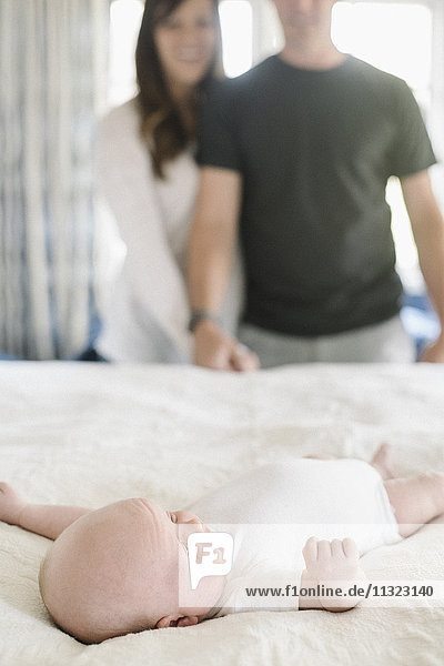 Two parents holding hands  standing over a young baby lying on a bed.