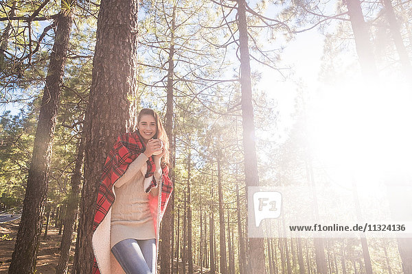 Lächelnde junge Frau mit Decke und Getränk an Baumstamm gelehnt im Wald