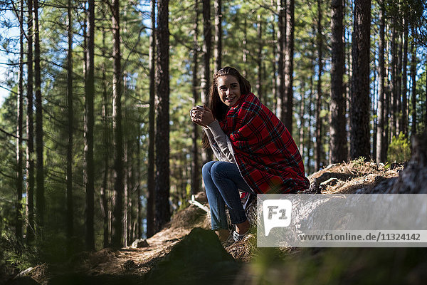 Lächelnde junge Frau mit Decke und Getränk auf Felsen im Wald sitzend