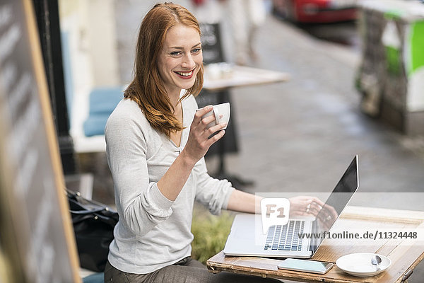 Rothaarige Frau mit Tasse Kaffee und Laptop im Straßencafé