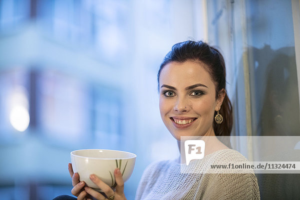 Porträt der lächelnden jungen Frau mit Teeschale vor dem Fenster