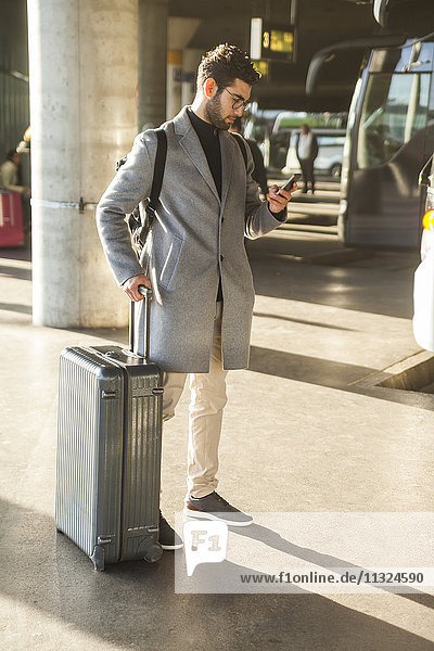 Businessman with baggage standing at bus terminal looking at cell phone