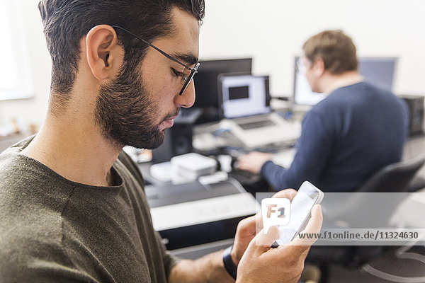 Young man using smartphone in modern office with coworker in background