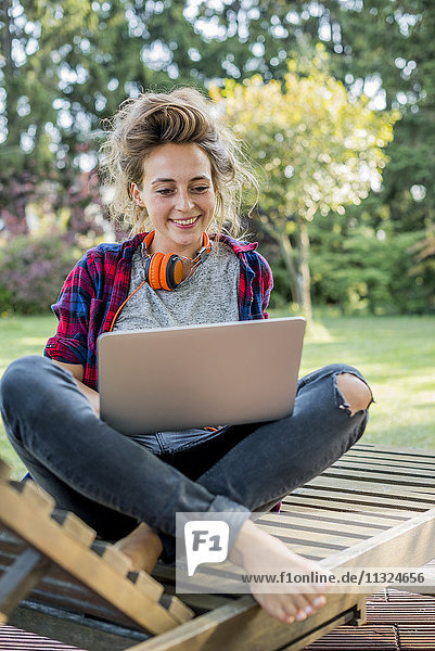 Porträt einer jungen Frau die auf einer Liege im Garten sitzt und einen Laptop benutzt.