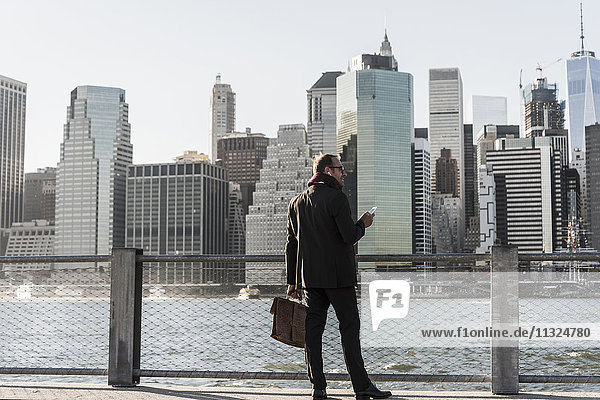 USA  Brooklyn  businessman with briefcase and smartphone standing in front of Manhattan skyline