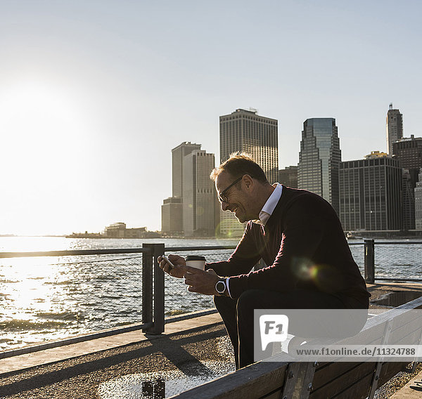 USA  Brooklyn  lächelnder Mann mit Kaffee zum Sitzen auf der Bank mit Blick auf Smartphone