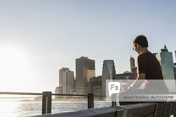 USA  Brooklyn  Frau mit Kaffee zum Sitzen auf der Bank mit Blick auf Smartphone