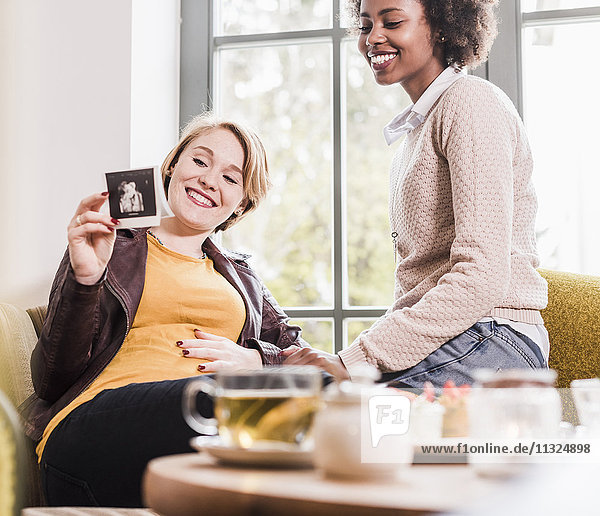 Pregnant young woman showing ultrasound scan to friend in a cafe