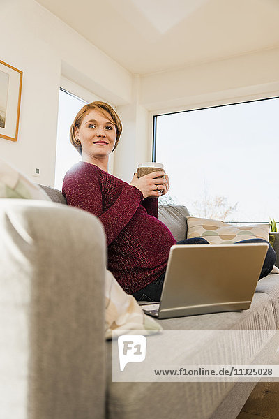 Schwangere Frau mit Laptop auf der Couch hält Tasse