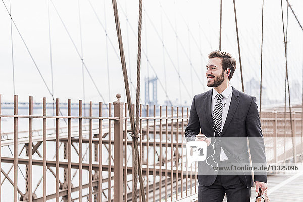 USA  New York City  smiling businessman with cell phone and earbuds on Brooklyn Bridge