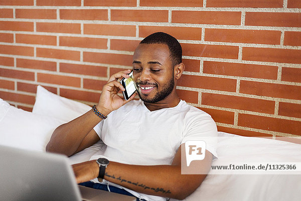 Lächelnder junger Mann im Bett mit Laptop und Handy