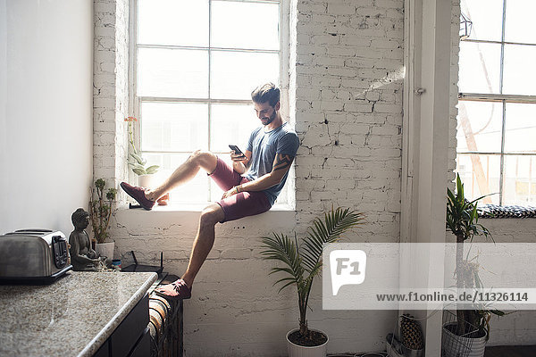Junger Mann sitzt auf der Fensterbank und schaut auf ein Handy in einem Loft.