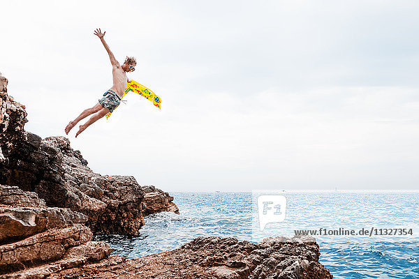Mann mit Luftmatratze springt vom Felsen ins Meer