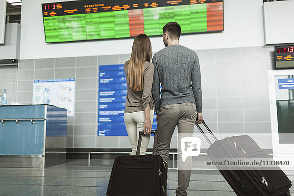 Rückansicht des jungen Paares beim Betrachten der Fluginformationstafel am Flughafen