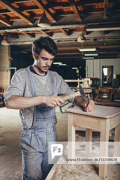 Carpenter hammering nail into wooden stool at workshop